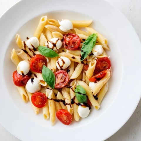 Close-up of vibrant Caprese Salad Pasta, featuring halved cherry tomatoes and creamy mozzarella pearls tossed with al dente fusilli and fresh basil leaves on a white ceramic plate.