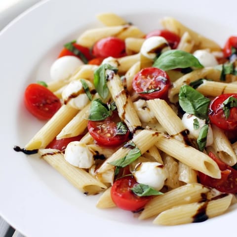 Overhead view of Caprese Salad Pasta, with glossy penne noodles, juicy tomato halves, and mozzarella pearls glistening with olive oil and balsamic glaze.