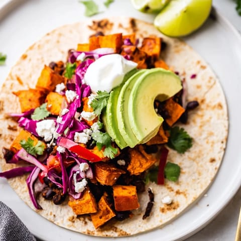 Golden roasted sweet potato and black bean tacos topped with avocado and cabbage, garnished with lime wedges on a rustic table.